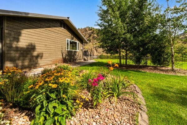 Side view of a brown house with a window, surrounded by a well-maintained yard featuring colorful flowers, green grass, trees, and gravel landscaping under a clear blue sky—ideal for recreational land with hills and water in the background.