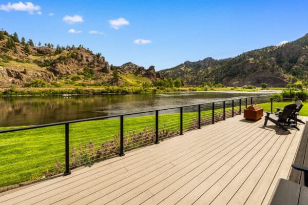 A wooden deck with black railing overlooks a calm river, green grass, and scenic rocky hills under a clear blue sky. Adirondack chairs and a fire pit invite relaxation on this beautiful recreational land or hunting property.