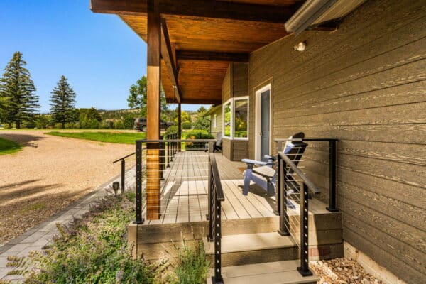 A sunlit porch with a wooden roof, cable railing, and two blue chairs overlooks a gravel driveway and green lawn with trees—perfect for enjoying the serene views of recreational land on a clear day.