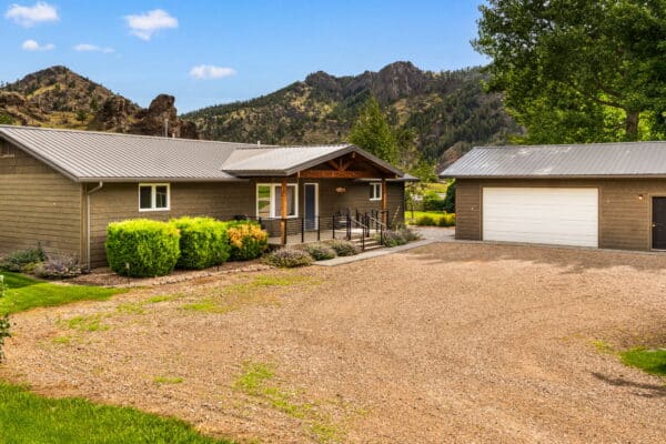 Single-story house with a metal roof, front porch, and ramp, surrounded by green bushes and a gravel driveway next to a detached garage. Set on recreational land with mountain views and blue sky in the background.