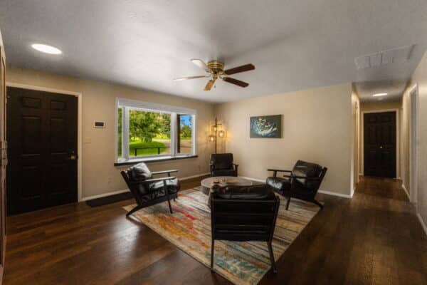 A cozy living room with dark wood floors, black leather chairs around a circular coffee table on a colorful rug, ceiling fan, large window with a green outdoor view ideal for recreational land, and wall art above a lamp.