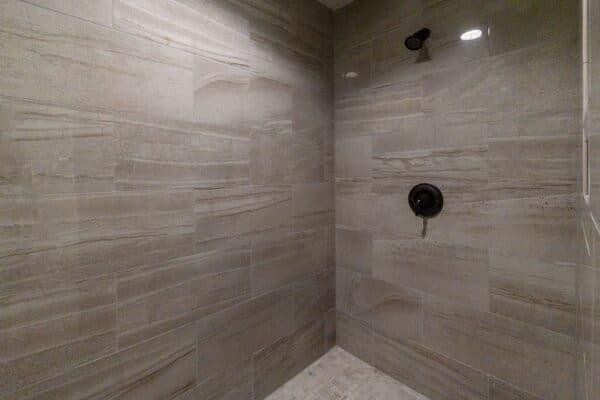 A tiled shower with large rectangular beige and brown tiles on the walls, a dark showerhead and faucet, and a light-colored small-tile floor—perfect for relaxing after exploring your new ranch for sale or hunting property. No shower curtain or door is visible.