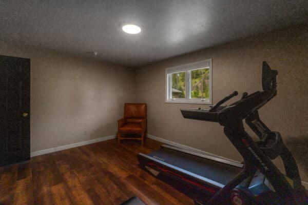 A small home gym room with wood flooring, beige walls, a window overlooking a cattle ranch, a single brown armchair, and a treadmill positioned beside the window. Natural light enters through the window.
