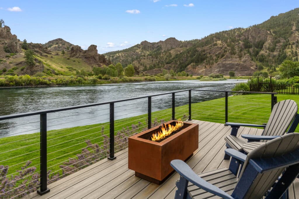 Deck with Adirondack chairs and a rectangular fire pit, overlooking recreational land with a grassy yard, calm river, and scenic, tree-covered hills under a clear sky—perfect for those seeking land for sale.