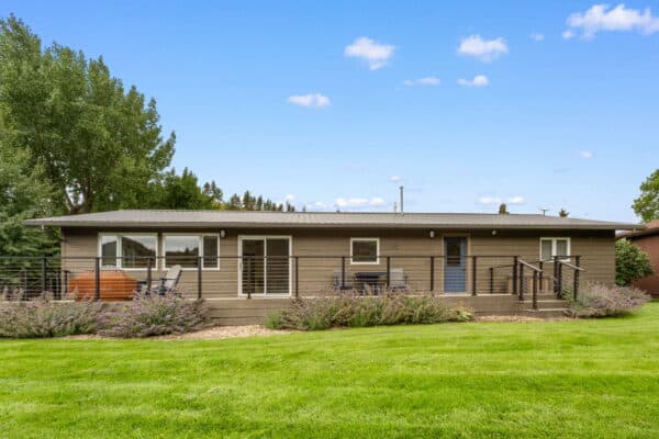 A single-story house with taupe siding, a black railing around the front porch, outdoor seating, and a well-kept lawn sits on recreational land surrounded by green trees under a blue sky with scattered clouds.