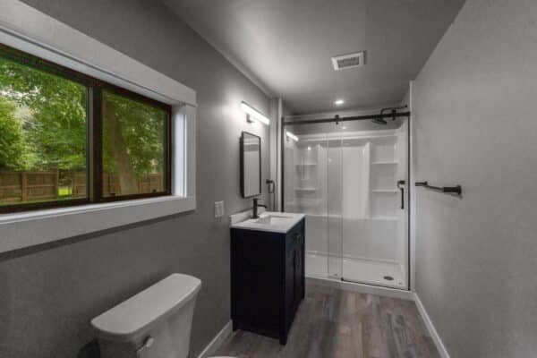 Modern bathroom with gray walls, a window overlooking trees, a white toilet, black vanity with sink and mirror, and a glass-enclosed shower on wood-patterned flooring—perfect for your hunting property or cattle ranch land for sale.