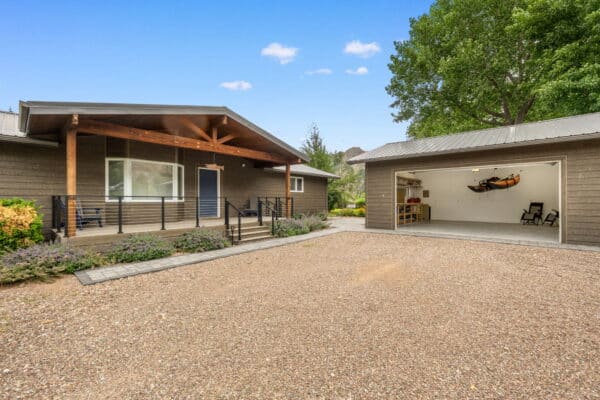 A modern single-story house on recreational land with a covered front porch, dark siding, and a detached garage with an open door. Inside the garage are shelves and a kayak. The yard features gravel and landscaping with shrubs.