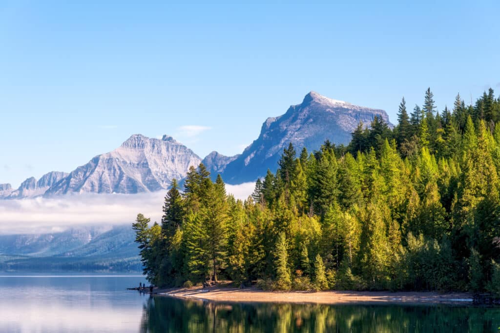 A calm lake reflects a forested shoreline with pine trees. In the background, snow-capped mountains rise under a clear blue sky, making it an ideal recreational land or hunting property for nature enthusiasts.