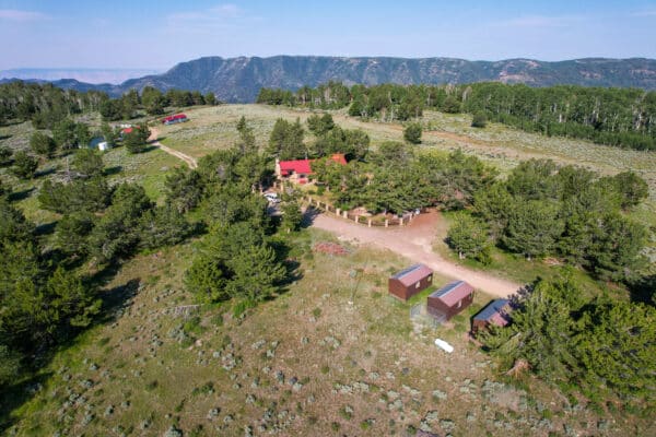 Aerial view of a rural cattle ranch with several small buildings and cabins surrounded by trees, open fields, and mountains in the background under a blue sky.