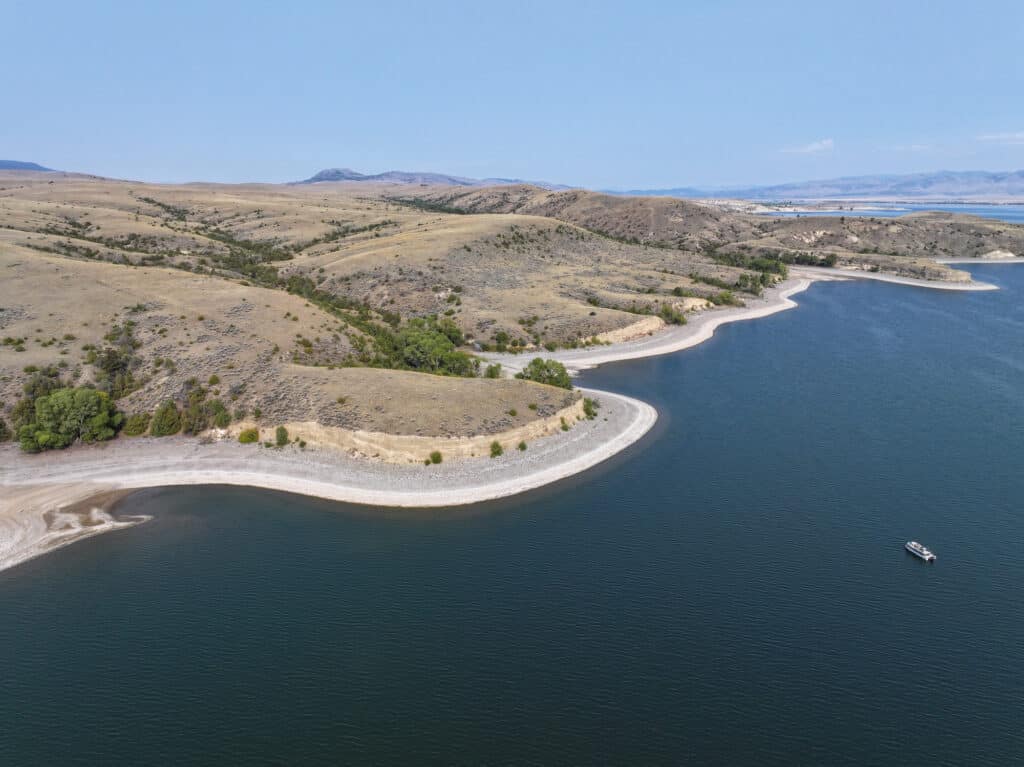 Aerial view of recreational land featuring a calm lake with a sandy, curving shoreline and rolling, grassy hills. Sparse vegetation and a small boat are visible on the water under a clear blue sky.