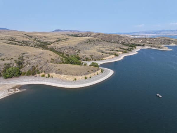 Aerial view of recreational land featuring a calm lake with a sandy, curving shoreline and rolling, grassy hills. Sparse vegetation and a small boat are visible on the water under a clear blue sky.