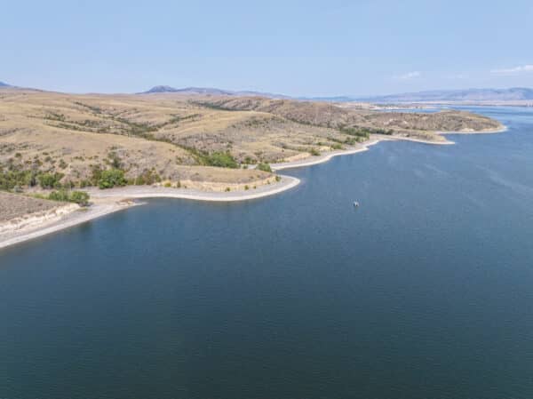 Aerial view of a wide, calm lake bordered by gently rolling, grassy hills and sparse trees under a clear sky. The shoreline curves smoothly around the water—an ideal setting for a cattle ranch or prime hunting property.