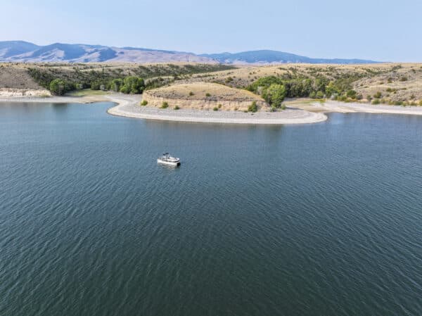 A lone boat floats on a calm lake near a curved shoreline with trees and grassy hills—an ideal setting for recreational land or a tranquil cattle ranch under a clear sky.
