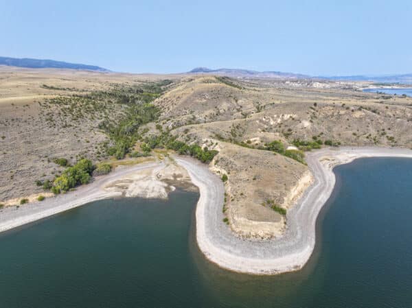 Aerial view of a curved, rocky shoreline surrounded by calm blue water and dry, rolling hills with sparse vegetation under a clear sky—ideal recreational land or cattle ranch land for sale.