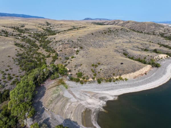 Aerial view of a lakeshore with a curved, pebbly beach, sparse trees, and dry, rolling hills under a clear blue sky. This scenic recreational land features an arid landscape with patches of green vegetation scattered throughout.