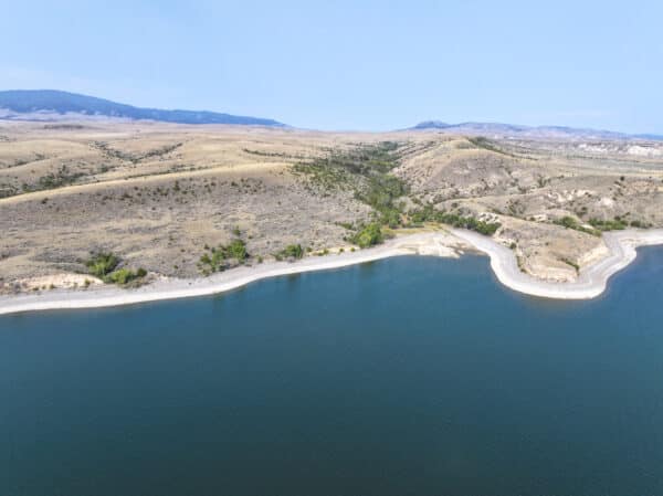 Aerial view of a calm lake with clear blue water bordered by a curved, rocky shoreline and dry, rolling hills—ideal for recreational land or as a peaceful hunting property under a clear sky.