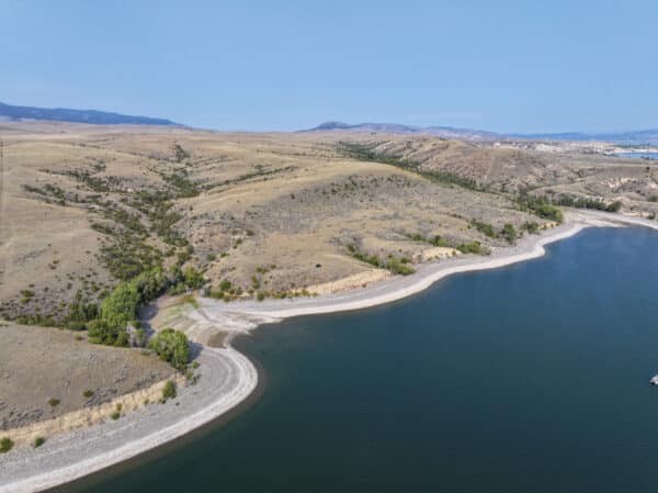 Aerial view of a calm lake with a rocky shoreline, nestled among rolling, dry hills with sparse vegetation—ideal recreational land or potential cattle ranch under a clear blue sky.