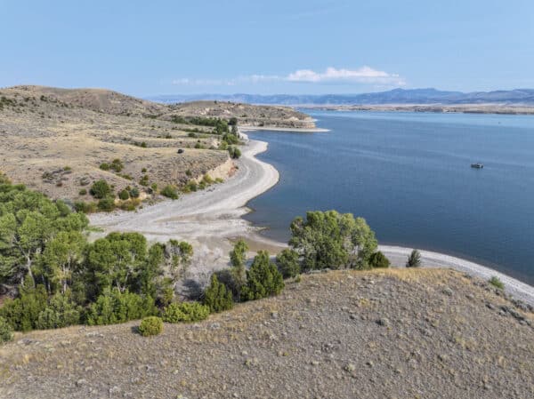 Aerial view of recreational land featuring a lake with a curving shoreline, surrounded by dry, grassy hills and patches of green trees under a clear blue sky. Mountains rise in the distance, and a small boat floats on the water.