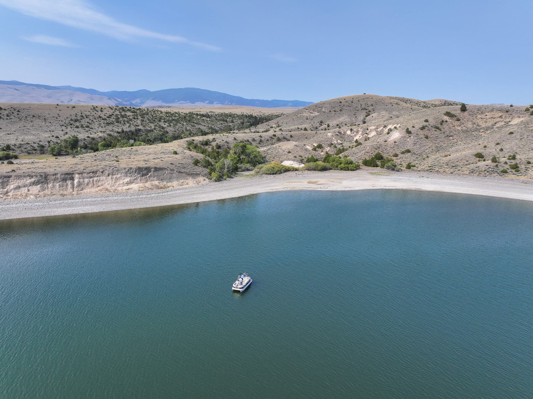 A small boat floats on a calm, blue lake near a rocky shoreline with gentle hills and scattered trees—an ideal view for a nearby cattle ranch or hunting property under the clear sky.