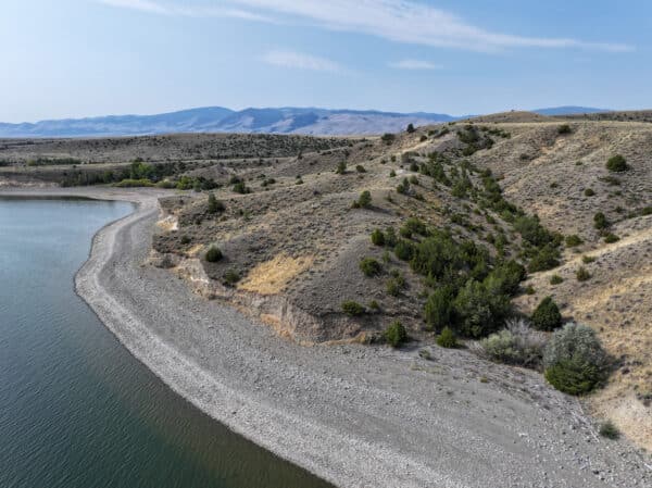 A wide, rocky shoreline curves along a calm body of water, bordered by dry, grassy hills with scattered bushes and small trees under a mostly clear sky and distant mountains—ideal land for sale perfect for a cattle ranch or hunting property.