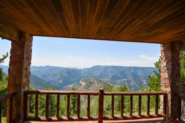 A wooden porch with a rustic railing overlooks a scenic mountain landscape on a picturesque cattle ranch. The porch roof and stone pillars frame the view, offering an inviting glimpse of this exceptional land for sale.