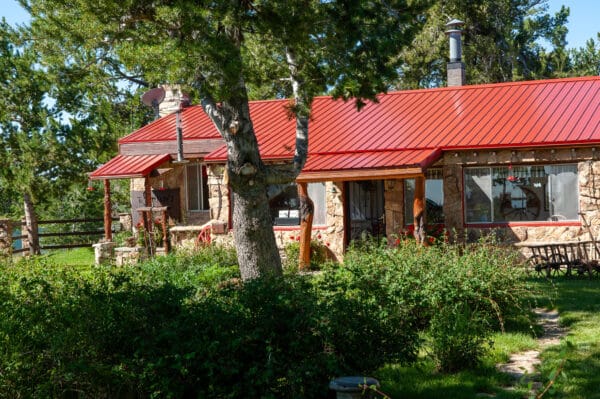 A rustic stone house with a red metal roof sits amid green trees and thick bushes on a sunny day. Perfect as a hunting property or ranch for sale, the yard is lush with grass and features a wooden bench near the house.