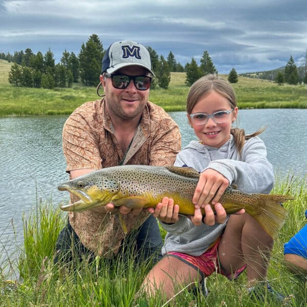 A man and a young girl kneel by a river, both smiling and holding a large brown trout together. Grassy banks, trees, and cloudy skies create an idyllic setting—perfect for enjoying the outdoors on your dream cattle ranch or ranch for sale.