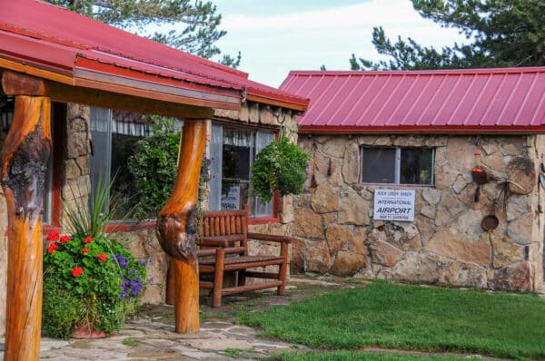 A rustic stone building with red metal roofs, wooden columns, a bench, hanging plants, colorful flowers, and a sign reading Rock Creek Ranch International Airport above a small window—perfect for a cattle ranch or recreational land. Grass and trees are visible.