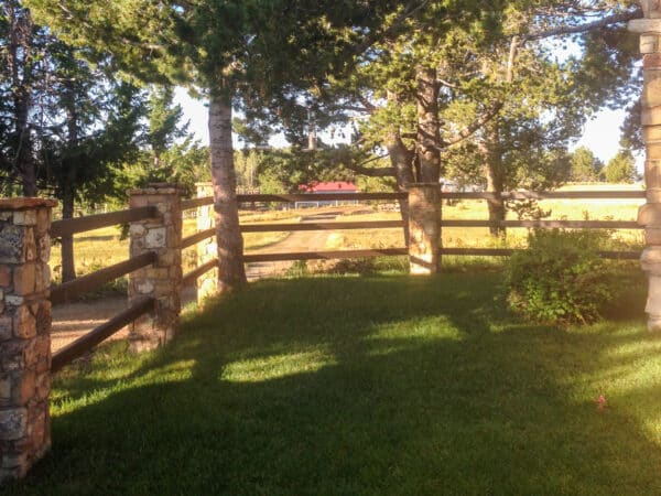 A sunlit, grassy yard on a recreational land is bordered by a wooden and stone fence, with tall trees casting shadows. In the background, a dirt road leads to a red-roofed building surrounded by fields and more trees.