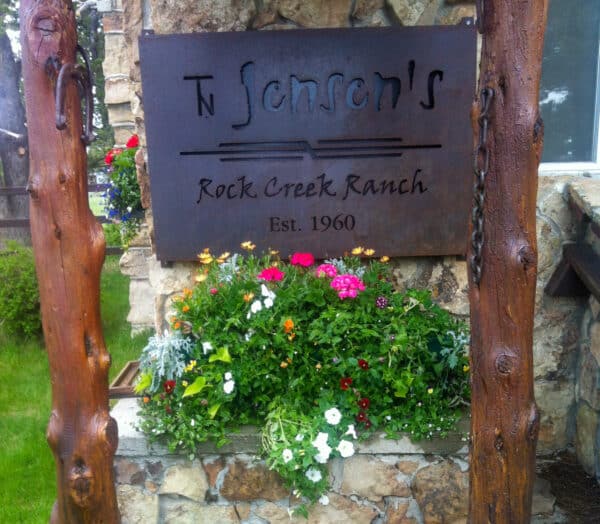 A rustic metal sign reading TN Jensenss Rock Creek Ranch Est. 1960 is mounted above a stone planter filled with colorful flowers, highlighting this scenic cattle ranch set against a stone wall and lush greenery.