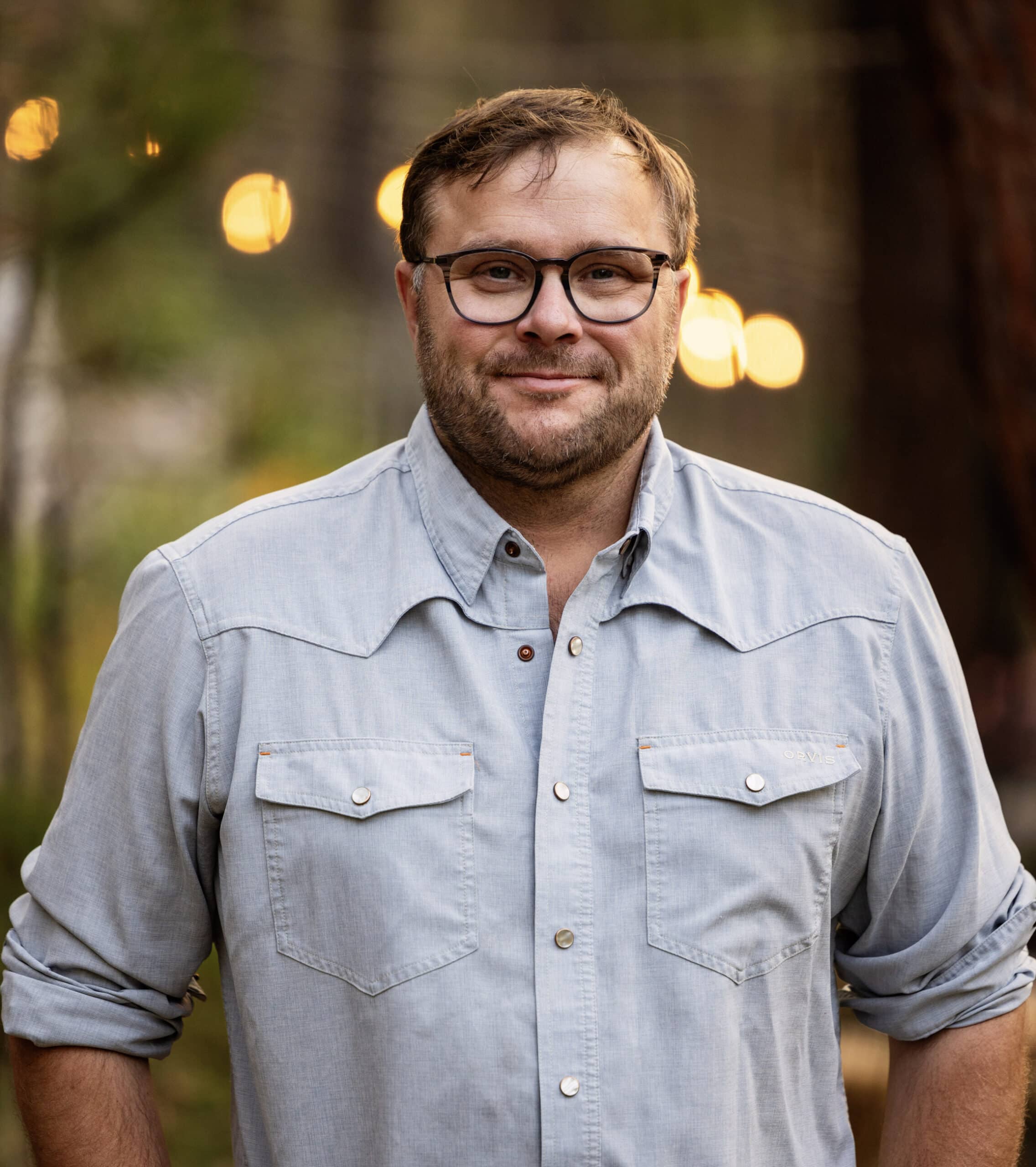 A man with short brown hair, glasses, and a beard is smiling while standing outdoors on recreational land. He is wearing a light blue button-up shirt with rolled-up sleeves. Blurred lights and trees are visible in the background.