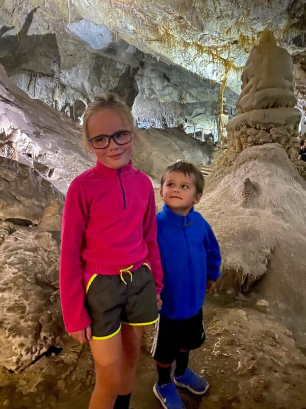 Two young children, a girl in a bright pink jacket and a boy in blue, stand inside a cave with rocky walls. The scene captures the adventure of exploring nature—ideal for families searching for a hunting property or ranch for sale.
