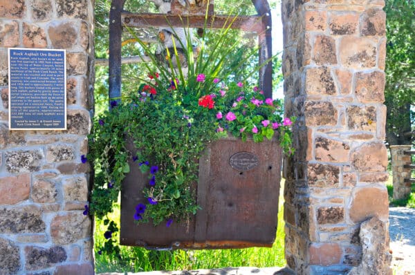 A large metal ore bucket filled with vibrant flowers hangs between two stone pillars, reminiscent of a historic cattle ranch. A plaque is mounted on the left pillar, as sunlight highlights the greenery and colorful blooms.