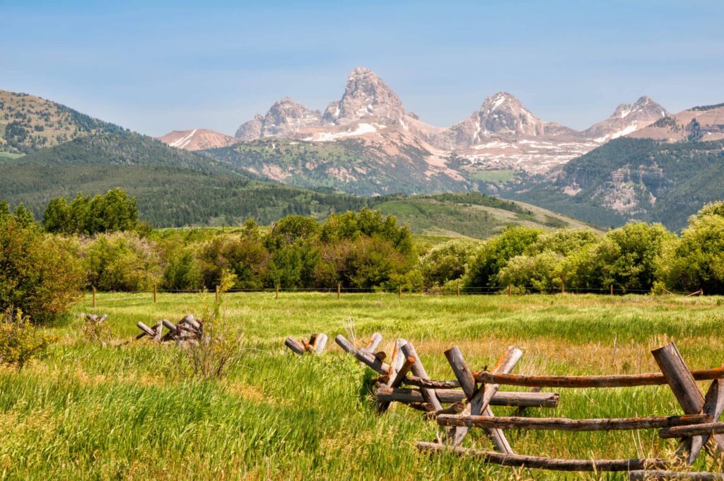 A rustic wooden fence zigzags through a green field with dense trees on recreational land, set against rolling hills and rugged, snow-capped mountain peaks beneath a clear blue sky.