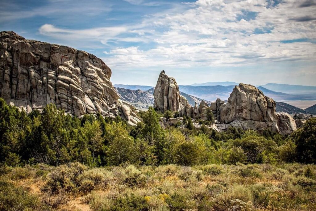 Large rocky formations rise above a landscape of green shrubs and trees under a partly cloudy blue sky, with distant mountains and a body of water visible—an ideal setting for a ranch for sale or cattle ranch.