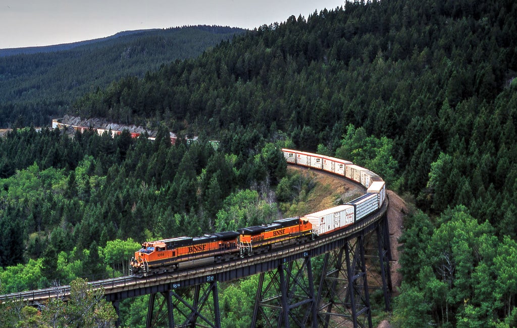 A long freight train with orange BNSF locomotives crosses a tall, curved bridge surrounded by dense green forest and rolling hills—an ideal setting for recreational land or a cattle ranch.