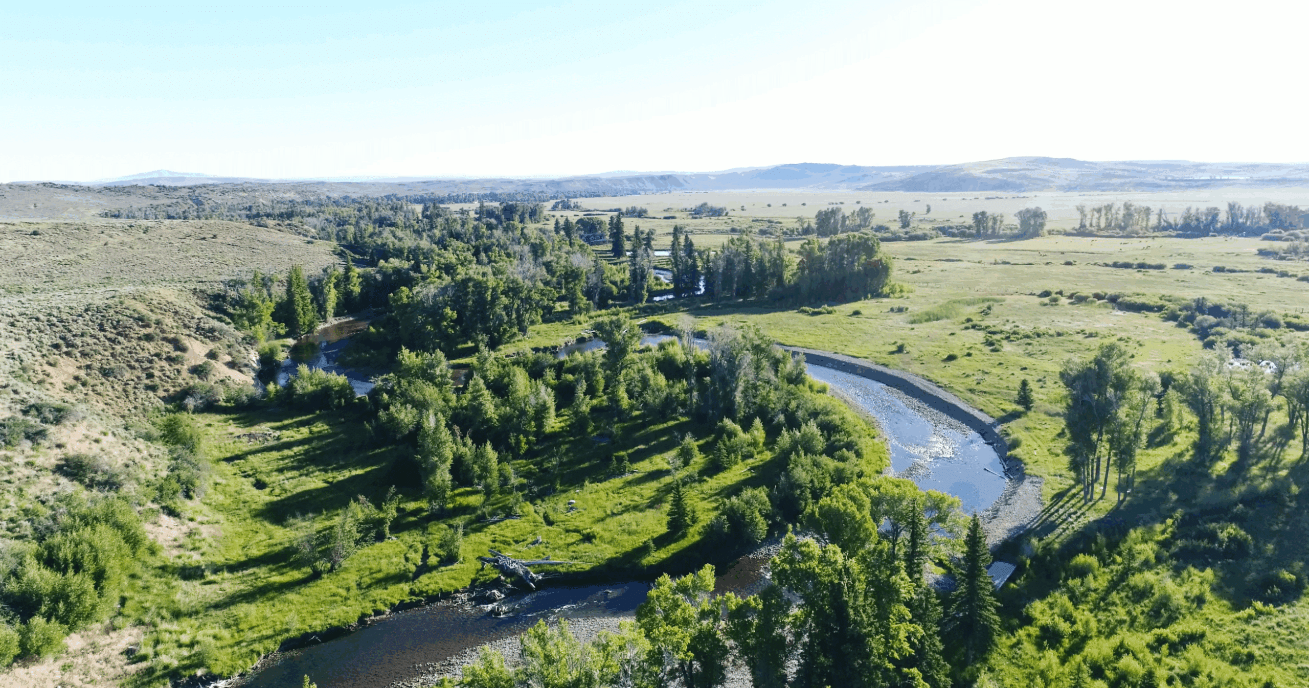 A winding river flows through a lush green valley with scattered trees and grassy meadows, bordered by low hills under a clear, bright sky—ideal recreational land or hunting property for those seeking land for sale.
