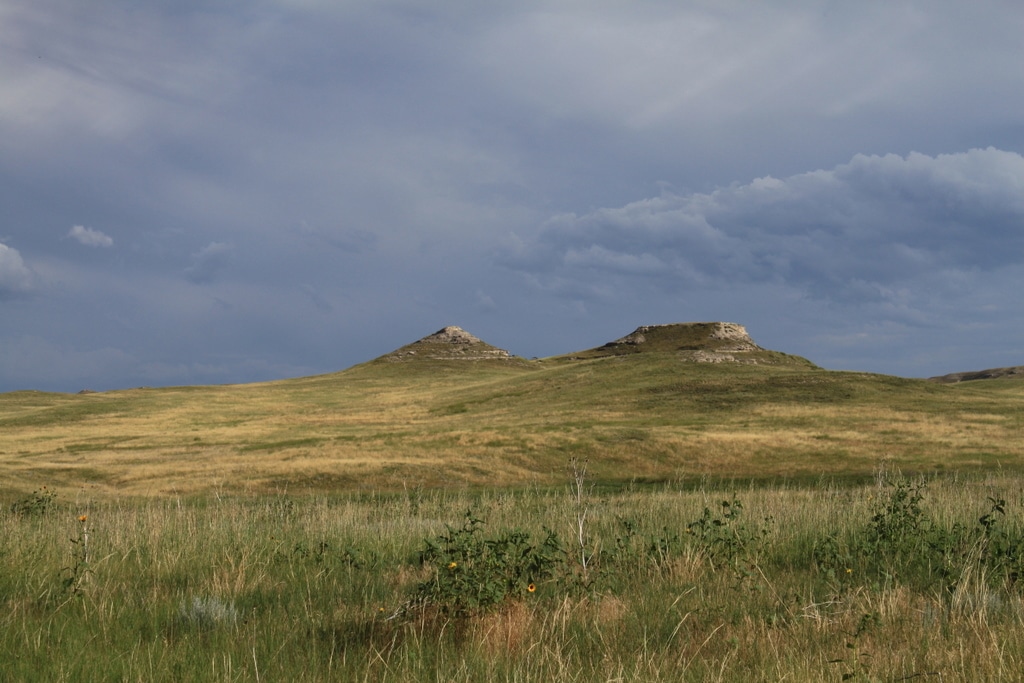 Grassy prairie landscape with two small, rocky buttes under a cloudy sky. Wildflowers and tall grasses are visible in the foreground—ideal recreational land or hunting property.
