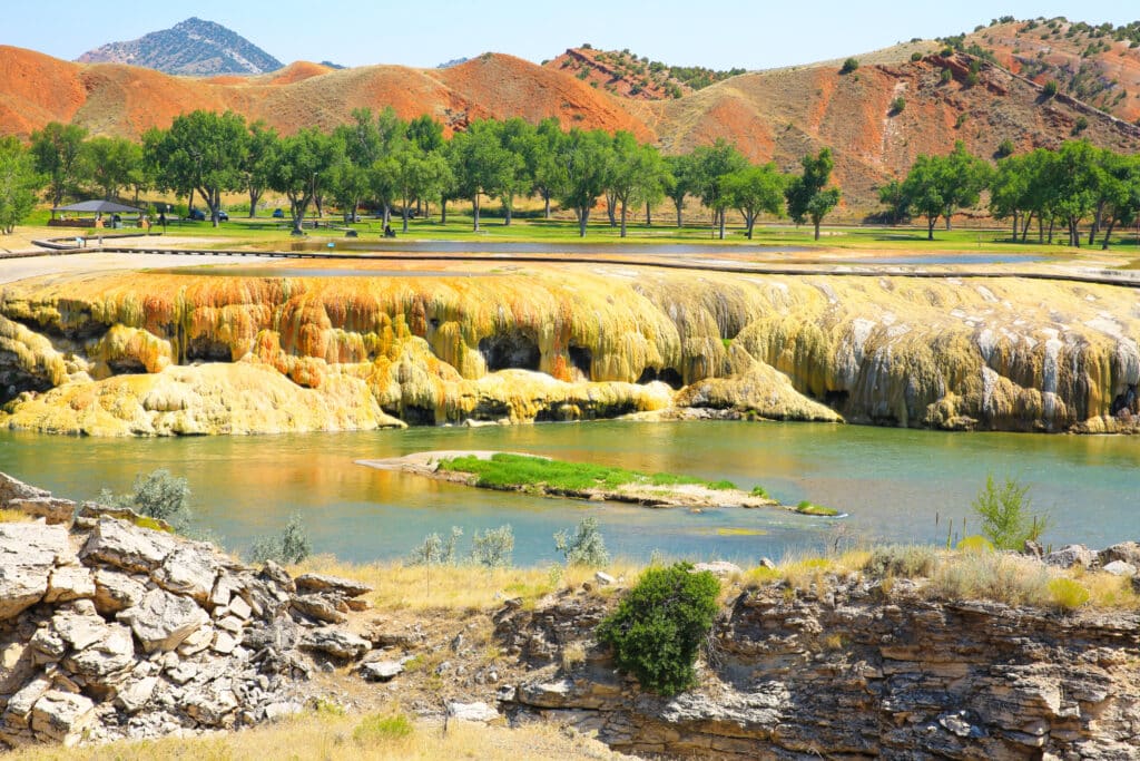 Colorful mineral terraces line a riverbank with green trees, grassy hills, and a distant mountain under a clear blue sky—picturesque recreational land in a scenic park setting.