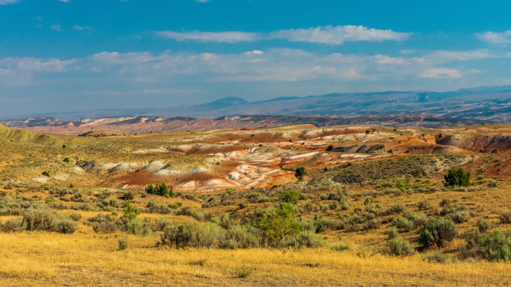 A wide desert landscape with rolling hills, patches of green bushes, and bands of red, yellow, and white earth under a blue sky with scattered clouds—distant mountains line the horizon. Ideal recreational land or ranch for sale.