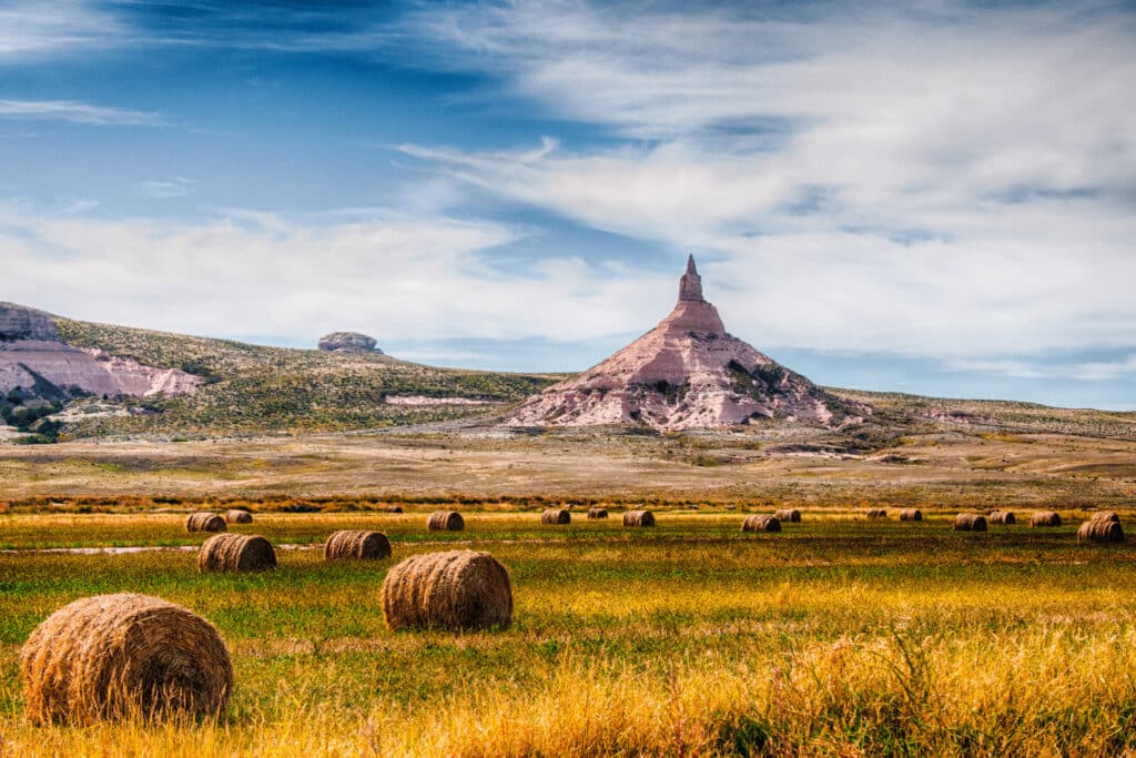 Hay bales scattered across a grassy field, with the distinctive Chimney Rock rising in the background under a partly cloudy blue sky—ideal recreational land or ranch for sale.
