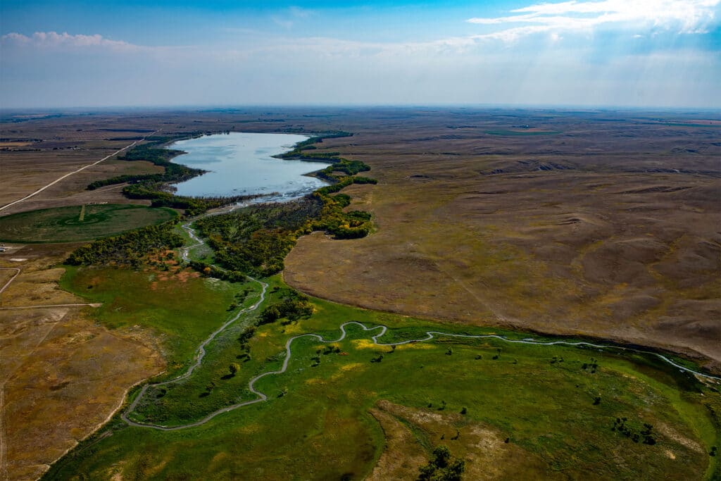 Aerial view of a winding creek leading to a large lake, surrounded by green vegetation and expansive brown grasslands under a blue sky—perfect recreational land or cattle ranch with scenic sunlight streaming through the clouds.