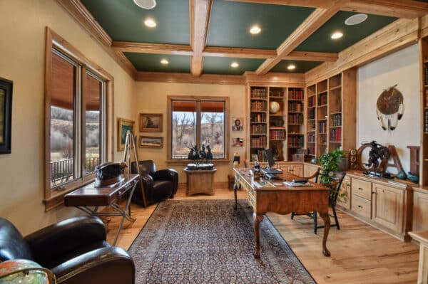 A spacious, rustic home office with wooden floors and beams, built-in bookshelves, a large ornate desk, leather chairs, Western decor, and windows overlooking scenic recreational land or a nearby cattle ranch.