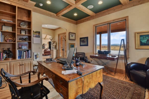 Home office with a wooden desk, two chairs, bookshelves, framed art, a computer monitor, and large window overlooking scenic recreational land and mountains. Warm, rustic decor and natural light fill the room.