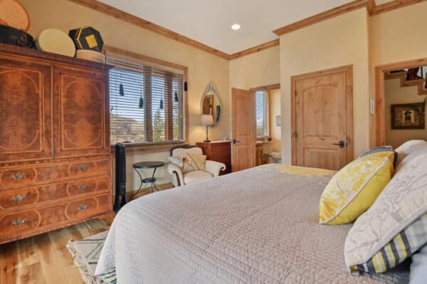 Bedroom with wood accents, ornate armoire, bed with patterned pillows, small writing desk by window with blinds, armchair with blanket, and doorway to a bathroom. Cozy atmosphere in neutral tones—perfect retreat on a cattle ranch or recreational land for sale.