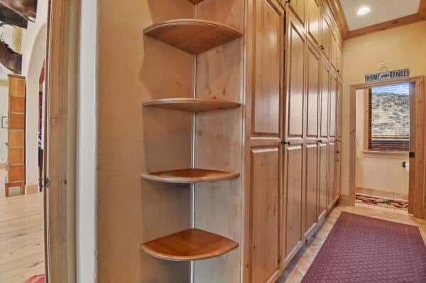 A hallway with wooden cabinets on the right, a corner shelf unit with three triangular shelves, and a maroon rug on a light wood floor. A window and a Home On The Range sign are visible—perfect for your dream ranch for sale.