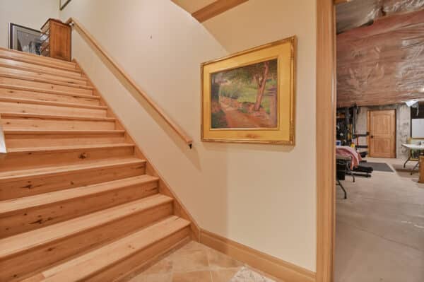 Wooden staircase with a smooth handrail leads up from a tiled floor. A framed landscape painting hangs on the beige wall. In the background, an unfinished basement evokes the rustic charm of a ranch for sale or recreational land.