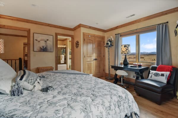 Cozy bedroom with patterned bedding, wooden trim, and a large window showcasing a scenic landscape. Perfect for a ranch for sale, the room features a desk, chair, armchair with red blanket, and a sheep picture on the wall.