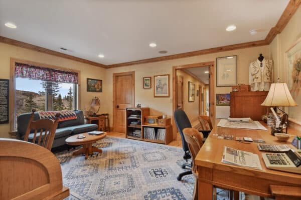 A cozy home office with wooden furniture, desk with papers and a lamp, chair, small blue sofa, carpeted floor, and large windows revealing trees and mountains—perfect for overseeing your recreational land or cattle ranch. Art and books decorate the room.