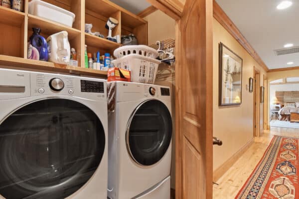 A laundry area with a front-loading washer and dryer, wooden shelves above holding detergents and a laundry basket. Ideal for a ranch for sale, the space opens into a hallway with wooden trim and a colorful rug leading to a bedroom.